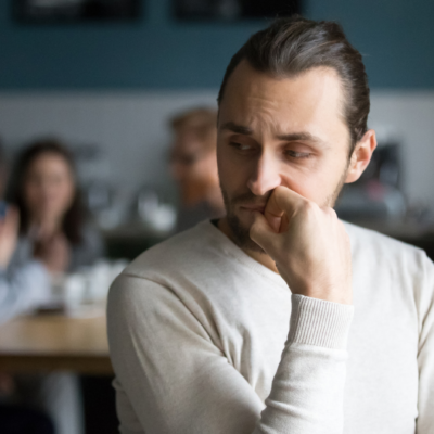 A man ponders with his face resting on his hand while friends mingle in the background.